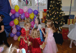 4 dziewczynki przebrane za księżniczki tańczą w kółku obok ramki z balonami i choinki